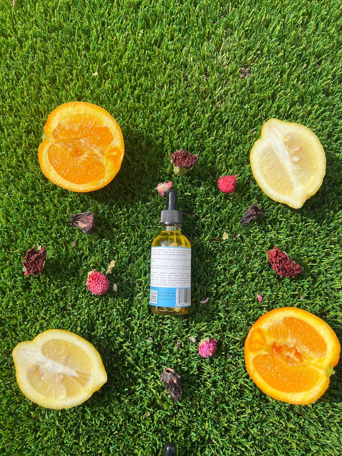 Bottle of Amor Sui Multi-Purpose Botanical Oil surrounded by orange and lemon halves on a grassy backdrop.