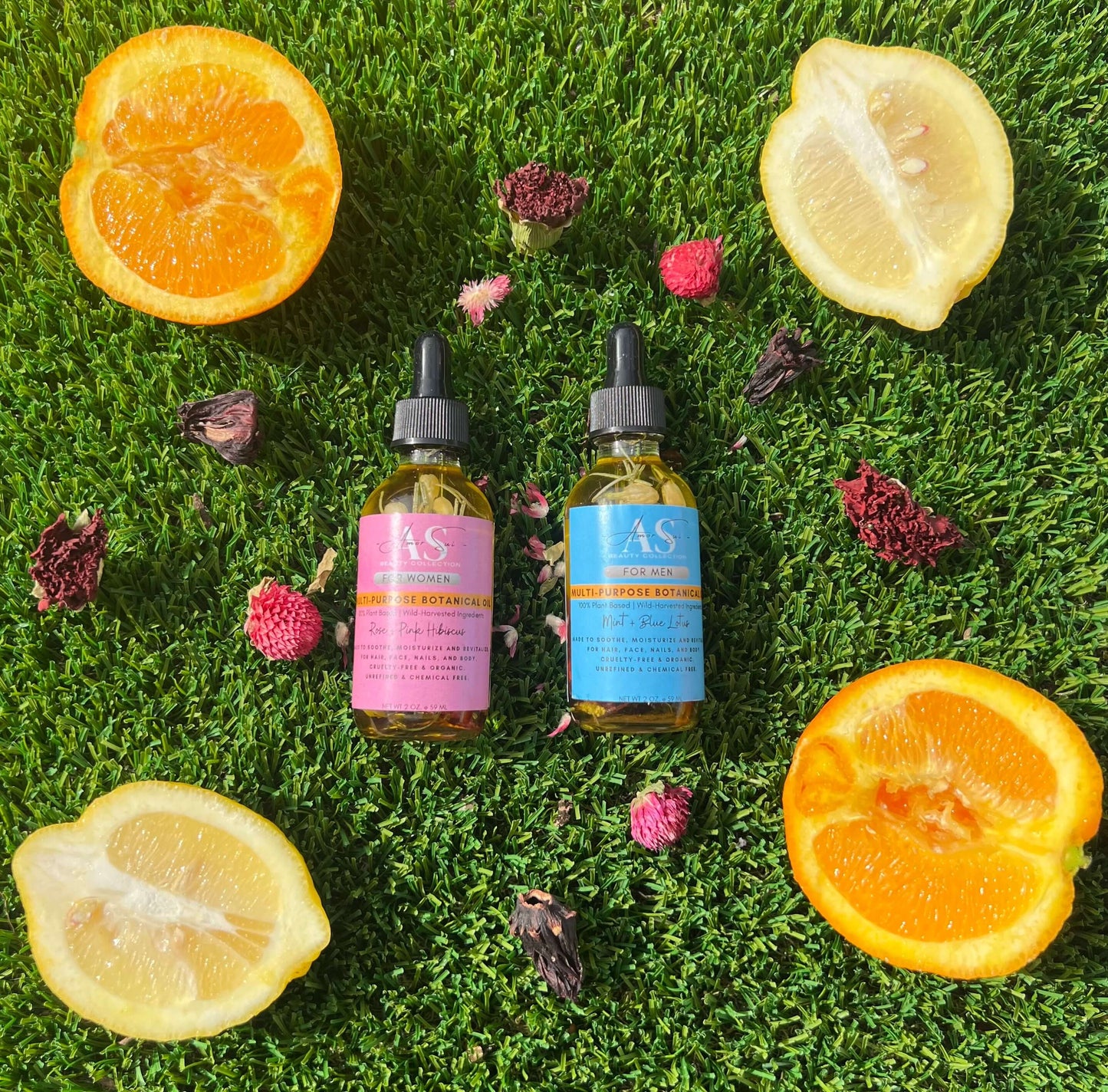 Two bottles of Amor Sui botanical oil on green grass, surrounded by orange and lemon slices, with flower petals.